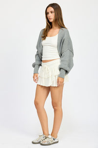 Woman wearing a gray cardigan, white top, and white skirt on a white background